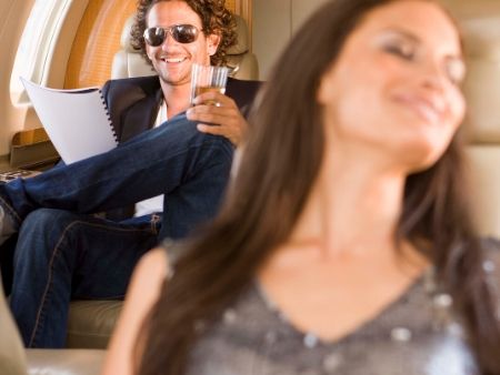 brunet sedí vzadu v lietadle a usmieva sa s pohárom vína v ruke na brunetu sediacu pred ním